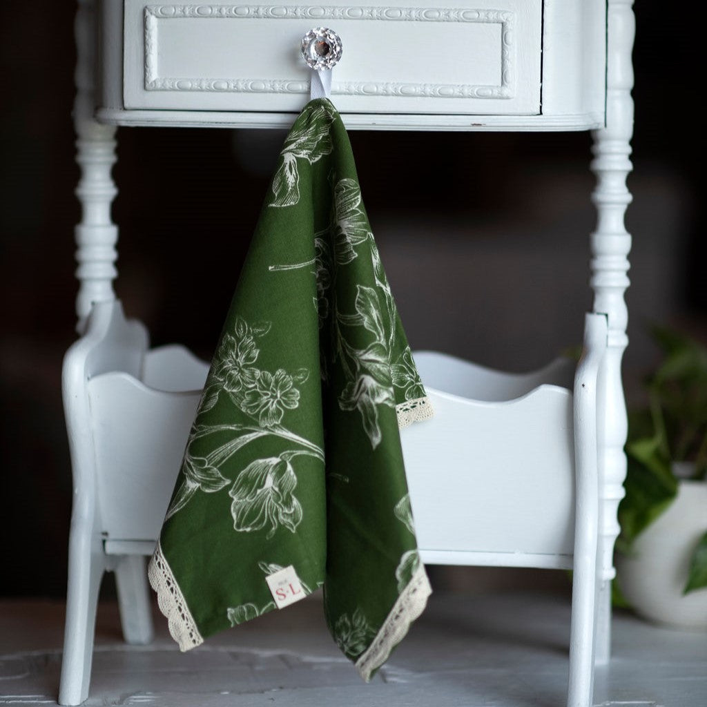 Linge à main de couleur vert olive avec fleurs blanches suspendu sur petit meuble en bois blanc.
