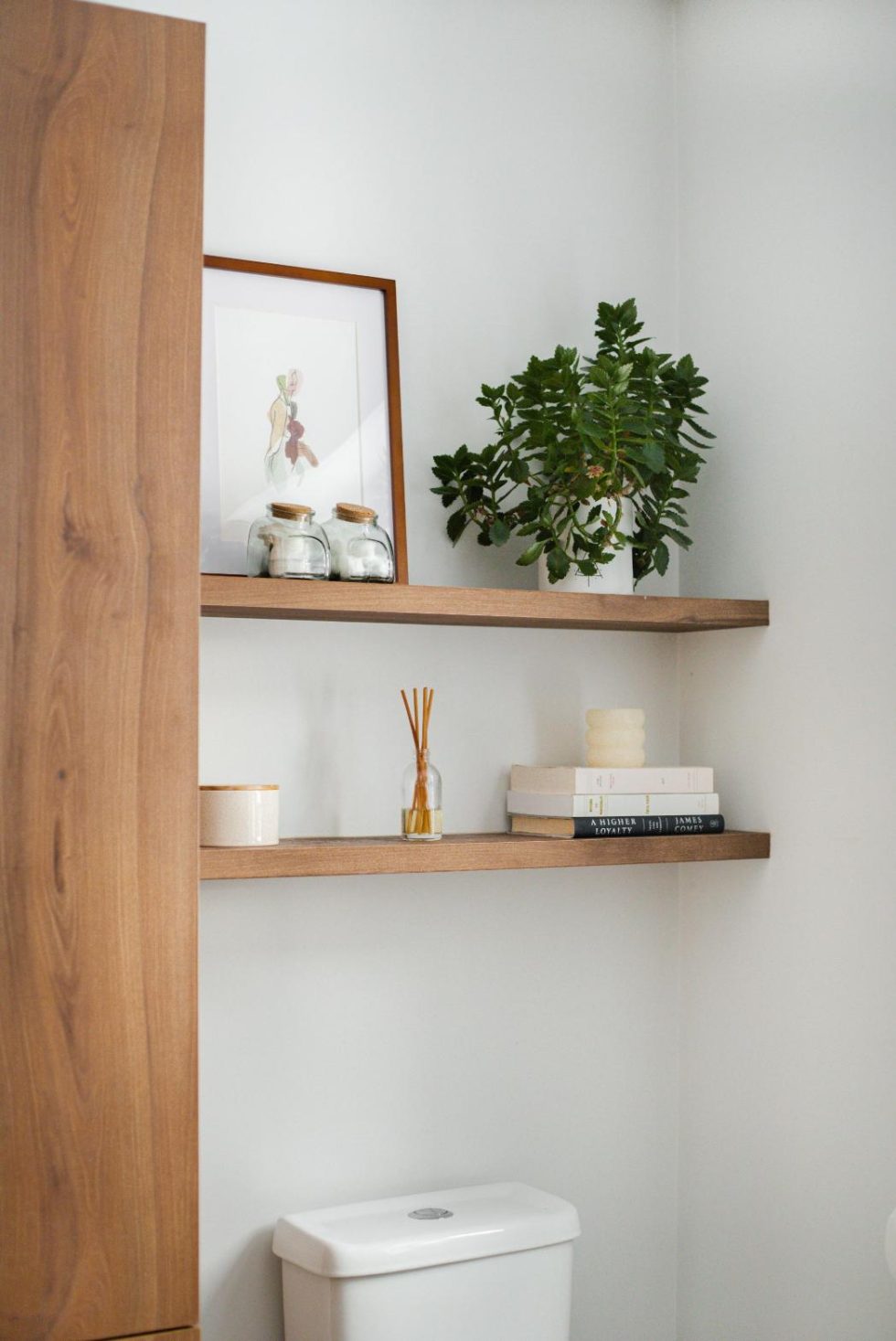 Salle de bain avec étagère en bois au-dessus des toilettes. Les étagères accueillent des plantes, des livres, des bocaux, un cadre photo et un diffuseur de parfum.
