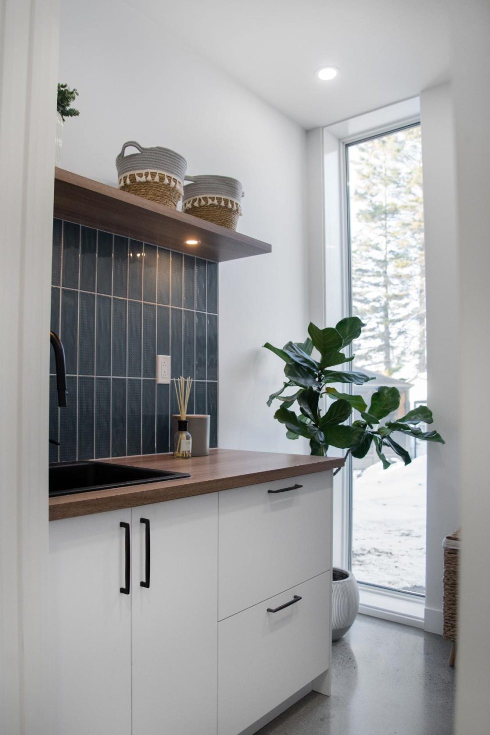 Coin cuisine minimaliste avec armoires blanches, comptoir en bois et dosseret en carrelage noir. Une grande plante se dresse près d'une grande fenêtre. Des paniers sont posés sur une étagère suspendue.