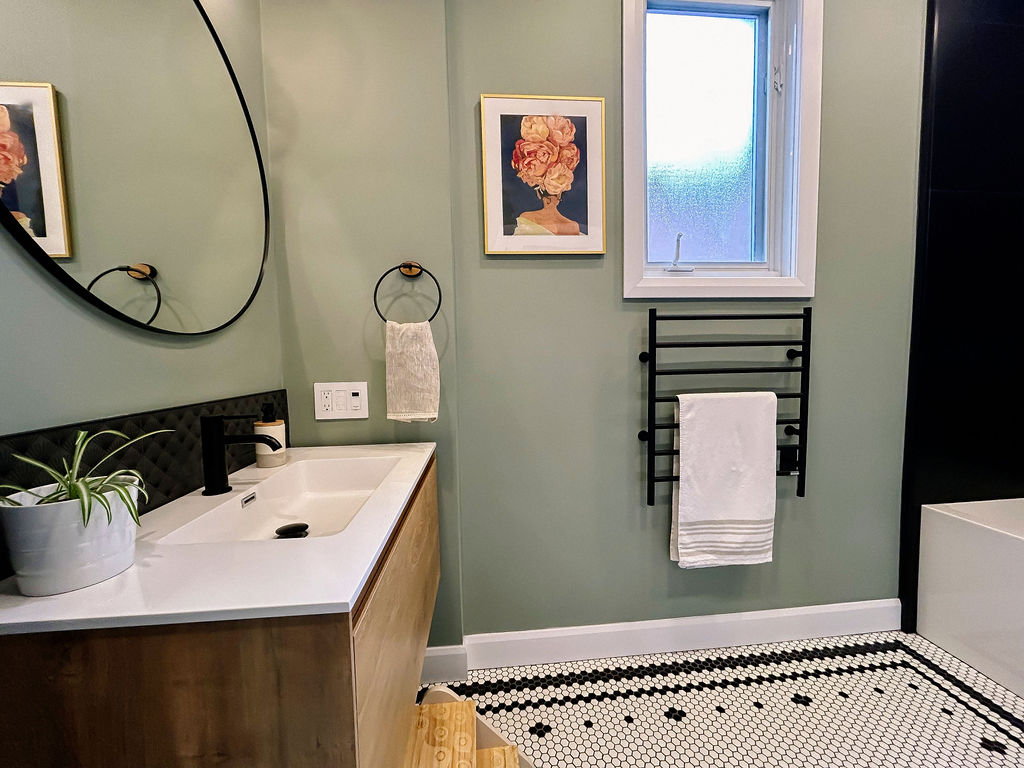 Salle de bain moderne avec meuble-lavabo en bois, lavabo blanc, miroir rond, porte-serviettes, cadre floral, murs verts et carrelage hexagonal noir et blanc. Une petite fenêtre se trouve au-dessus du porte-serviettes.