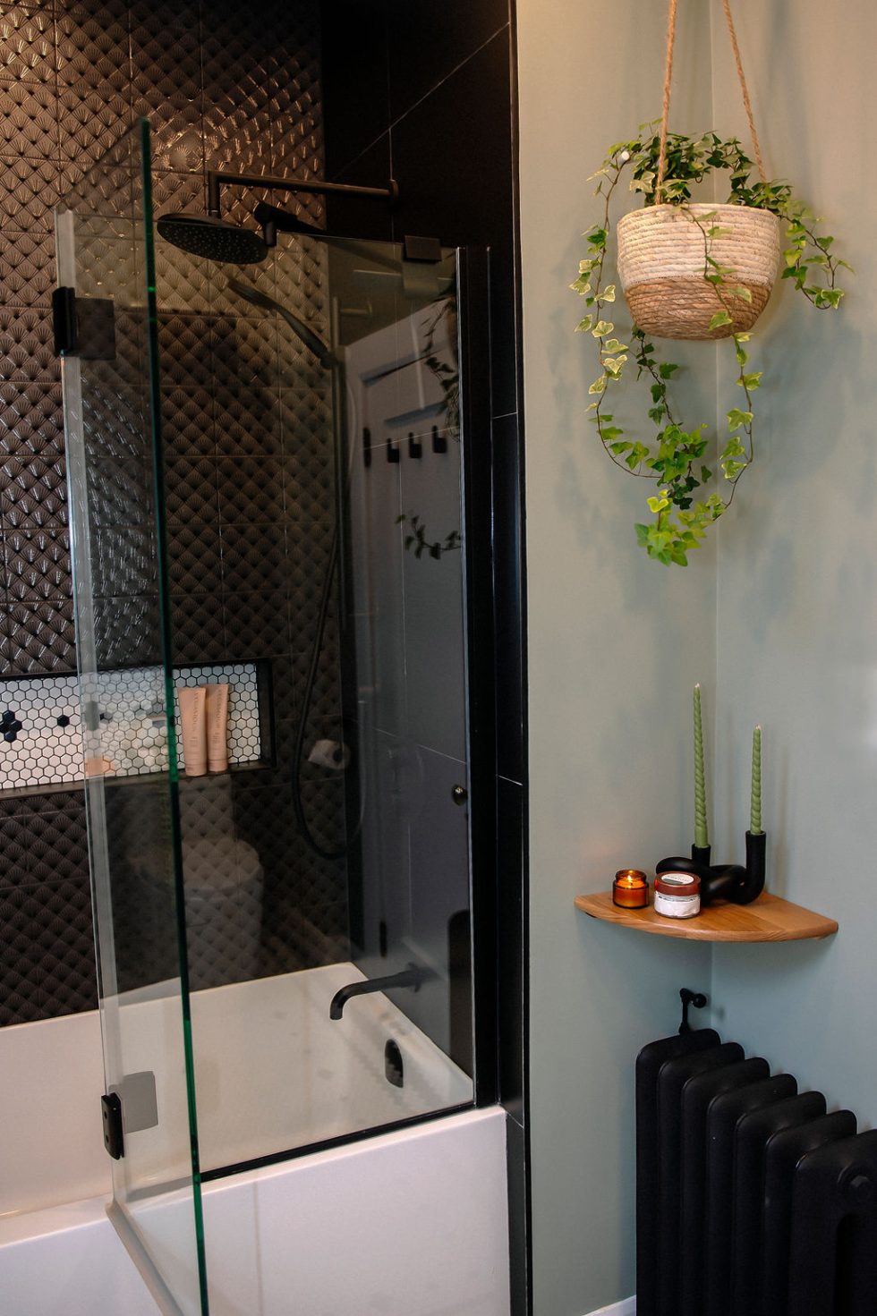Salle de bain moderne avec douche carrelée noire, porte vitrée, plante suspendue, étagère avec bougies et bougies vertes, et un radiateur noir.