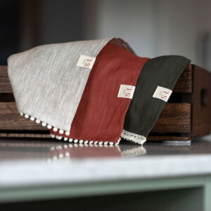 Trio de linges à main de couleurs naturelle, terre cuite et vert profond avec dentelle et pompons dans une boîte en bois sur comptoir.